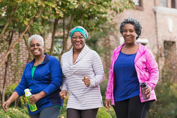 A group of three senior African America women exercising together, walking along a sidewalk in a residential neighborhood. They are having fun, smiling at the camera.